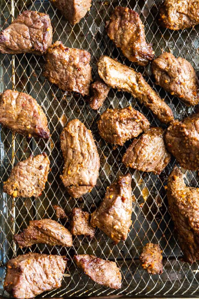 Close-up of cooked, marinated beef chunks on a wire mesh rack. The meat pieces are browned and textured, suggesting a grilled or roasted preparation. The background is a metal grid which enhances the rustic appearance of the dish.