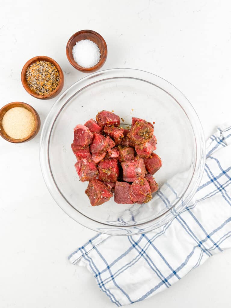 Chunks of raw seasoned meat in a glass bowl on a white surface. Three small bowls with salt, pepper, and spices are nearby. A white and blue checkered cloth is partially visible.