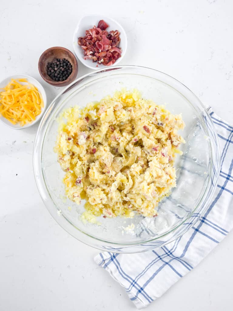 A glass bowl filled with mashed potatoes, bacon bits, and spices sits on a white countertop. Shredded cheese, a bowl of black pepper, and a small dish of bacon bits are nearby. A blue and white checkered cloth is placed beside the bowl.