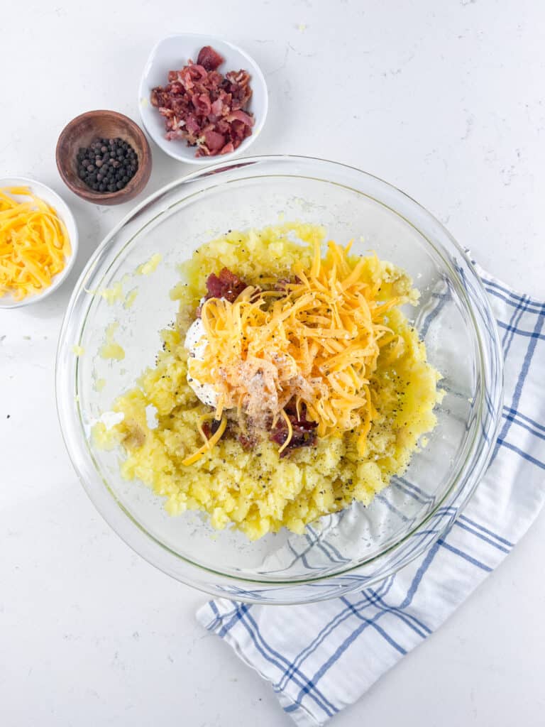 A glass bowl filled with mashed potatoes, shredded cheddar cheese, bacon bits, and a dollop of sour cream. Surrounding the bowl are small dishes containing more bacon, shredded cheese, and peppercorns, all on a white surface with a striped cloth.