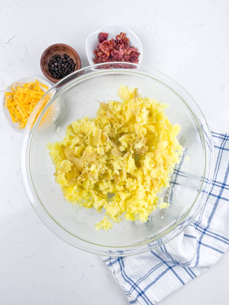 A glass bowl of mashed potatoes on a white countertop, surrounded by small bowls containing shredded cheese, bacon bits, and black pepper. A folded white and blue dish towel lies beside the bowl.