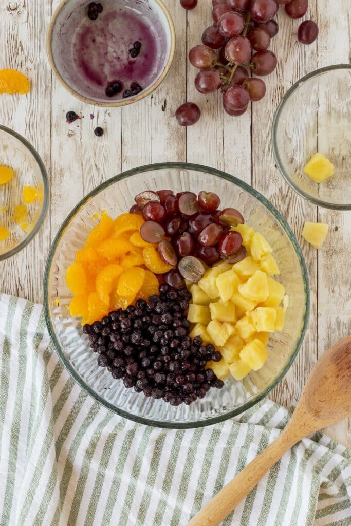 A glass bowl filled with chopped pineapples, grapes, blueberries, and mandarin segments on a wooden surface. Nearby are empty bowls with traces of fruit, a cluster of grapes, and a wooden spoon. A striped cloth is draped on the table.