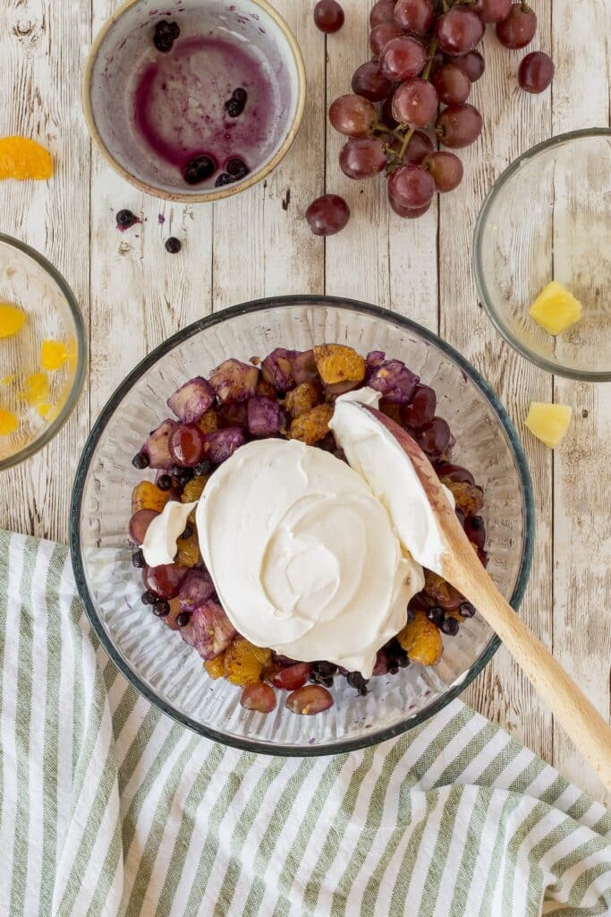 A glass bowl filled with grapes, berries, and orange slices, topped with whipped cream, and a wooden spoon. Surrounding the bowl are small bowls with fruit and juice, red grapes, and a striped cloth on a wooden surface.