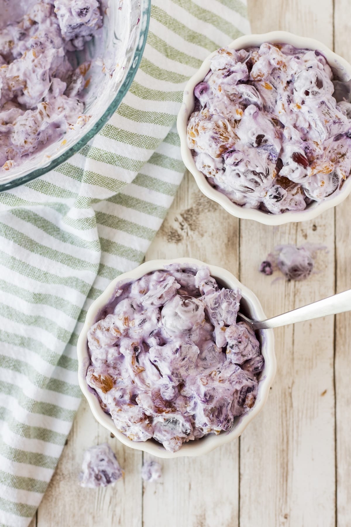 Two bowls of creamy, purple dessert with a spoon rest on a rustic wooden surface. A green and white striped cloth is partially visible nearby. The dessert appears to be a mixture of fruits and cream.