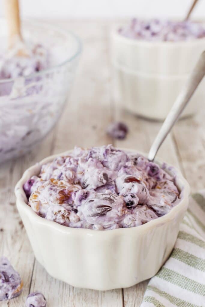 A bowl of creamy, purple-tinted fruit salad sits on a wooden surface. It contains grapes and nuts, mixed with a whipped topping. A spoon is placed in the bowl, and a striped cloth napkin is nearby, with another bowl in the background.