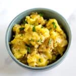 A bowl of creamy potato salad topped with chopped parsley, served in a light blue dish against a white background.