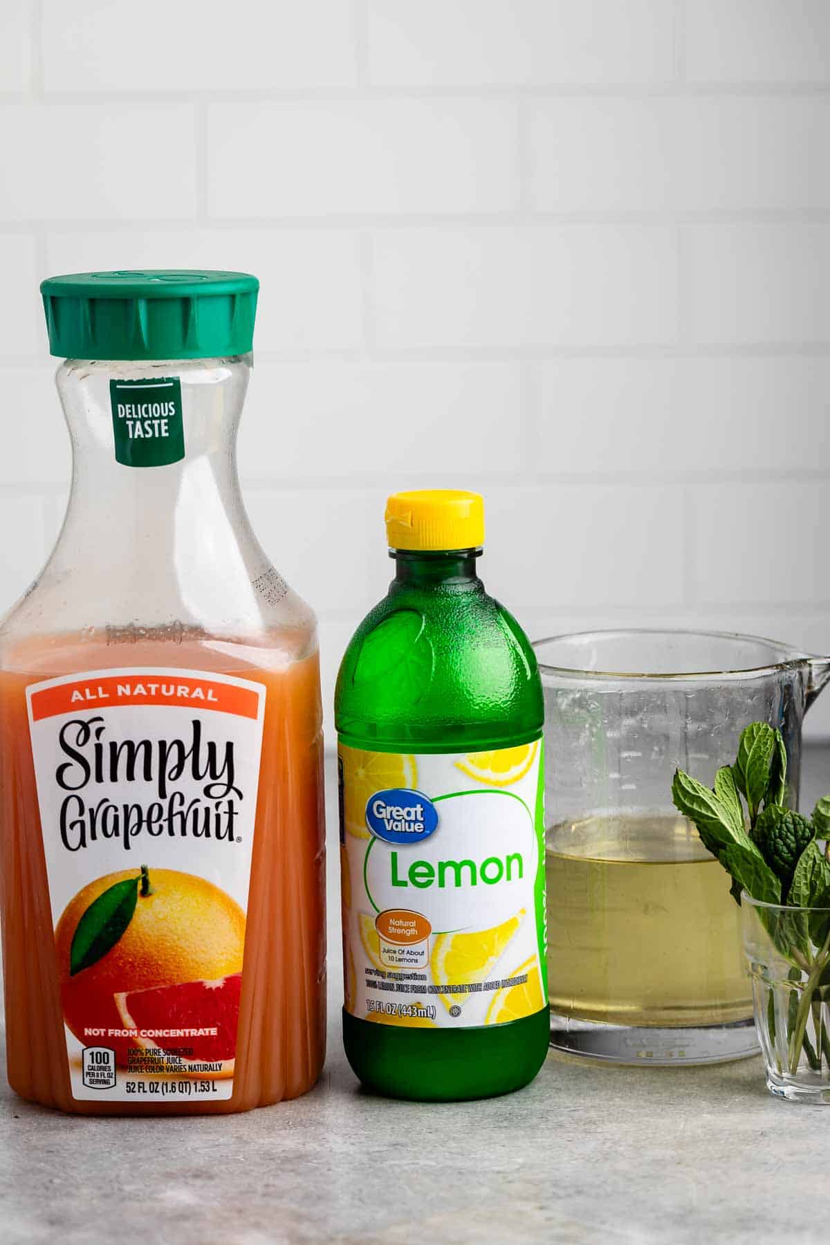 A bottle of Simply Grapefruit juice, a bottle of lemon juice, and a glass pitcher with a light liquid are on a countertop. Fresh mint leaves are next to the pitcher. The background features a white tiled wall.