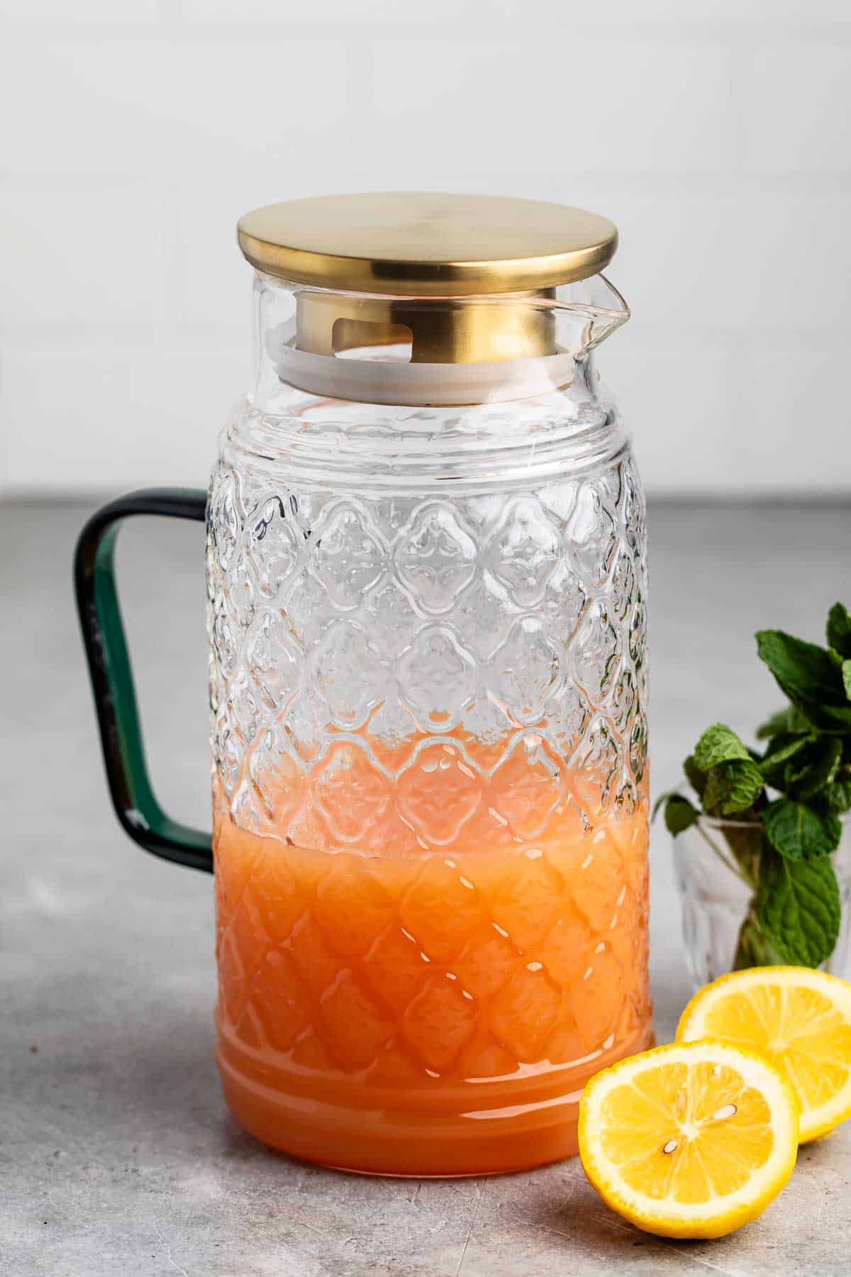 A textured glass pitcher filled halfway with orange liquid, topped with a golden lid. It has a dark green handle, and next to it are two lemon slices and fresh mint leaves on a gray surface. The background features a white tiled wall.