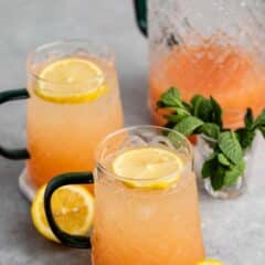 Two glass mugs filled with pink lemonade garnished with lemon slices. A clear pitcher in the background holds more of the beverage. Fresh mint leaves and lemon slices rest on the gray surface.