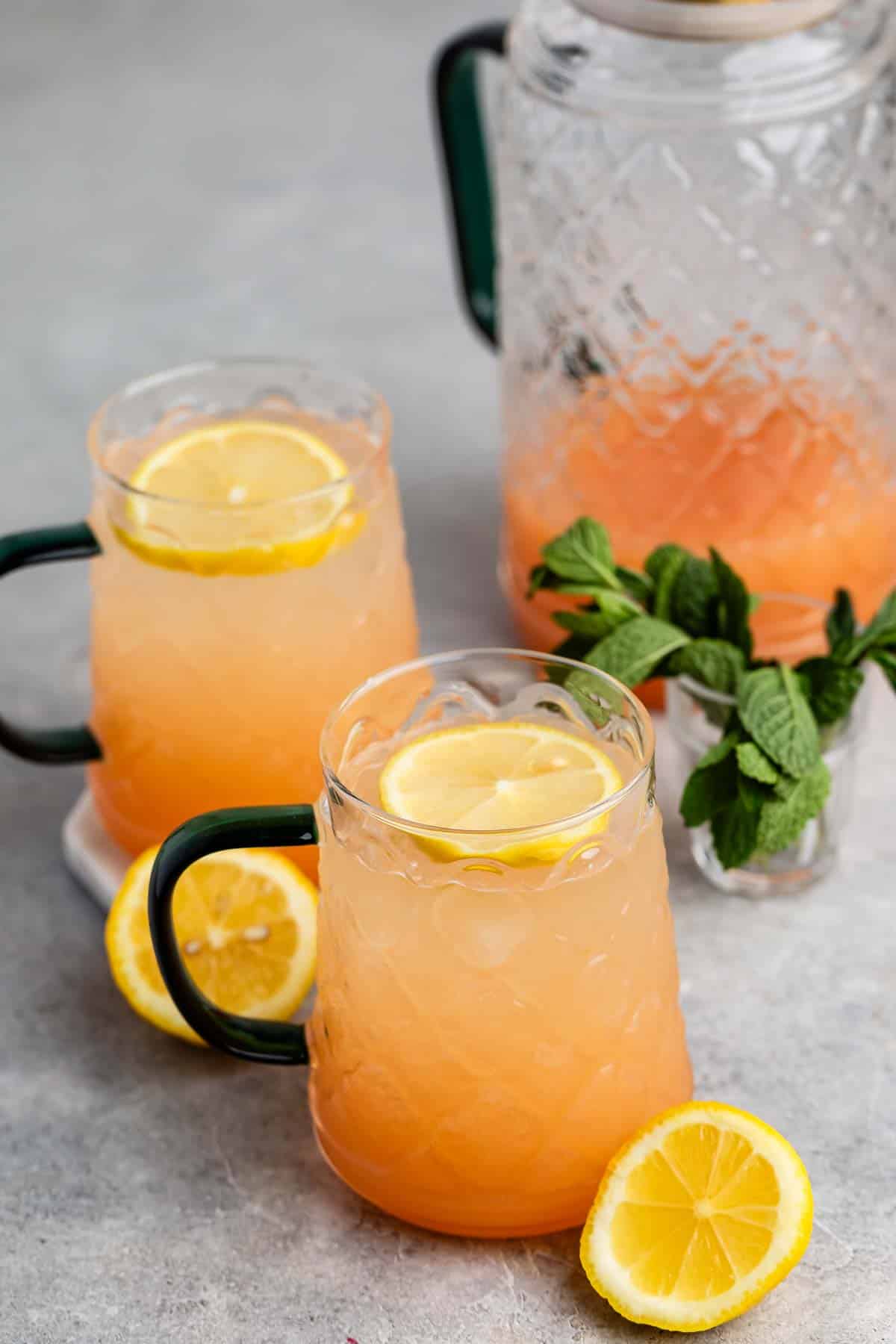 Two glass mugs filled with pink lemonade garnished with lemon slices. A clear pitcher in the background holds more of the beverage. Fresh mint leaves and lemon slices rest on the gray surface.