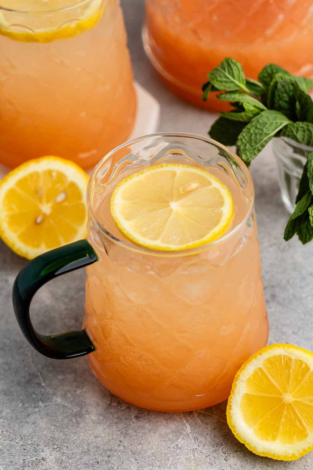 A glass mug filled with a peach-colored iced drink, garnished with a lemon slice on top. Fresh mint leaves and lemon slices are nearby on a gray surface, and additional similar drinks are in the background.