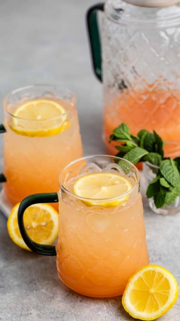 Two glass mugs filled with iced peach-colored lemonade garnished with lemon slices on a gray surface. A pitcher of the same drink and a bunch of fresh mint are in the background. A lemon slice is placed nearby.