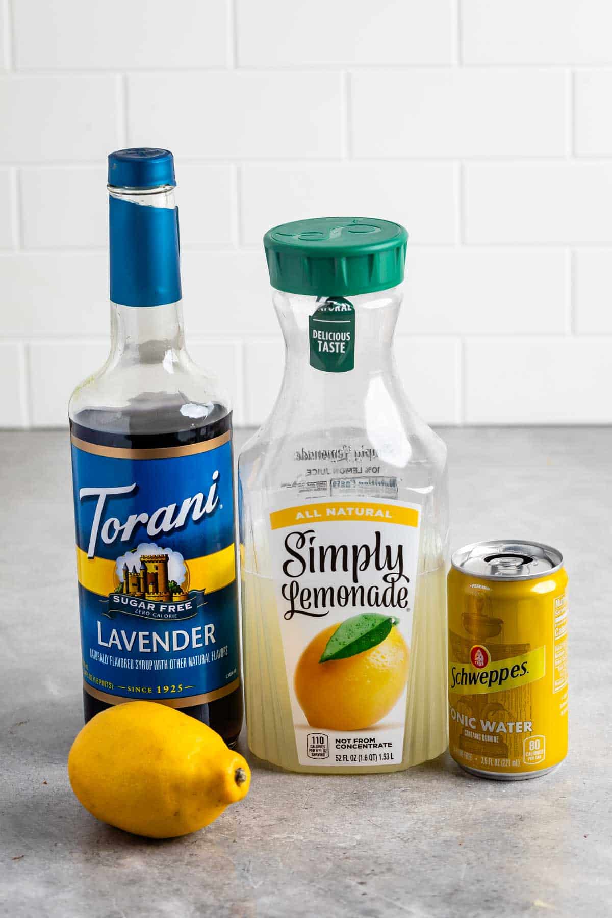 A bottle of Torani sugar-free lavender syrup, a Simply Lemonade container, a Schweppes tonic water can, and a lemon are arranged on a gray countertop with a white tiled background.