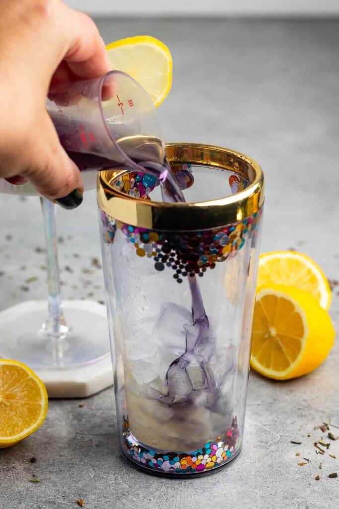 A hand pours a purple liquid from a small measuring cup into a glass filled with ice. The glass has a decorative pattern with colorful dots and a gold rim. Two lemon halves and a cocktail glass are in the background on a gray surface.