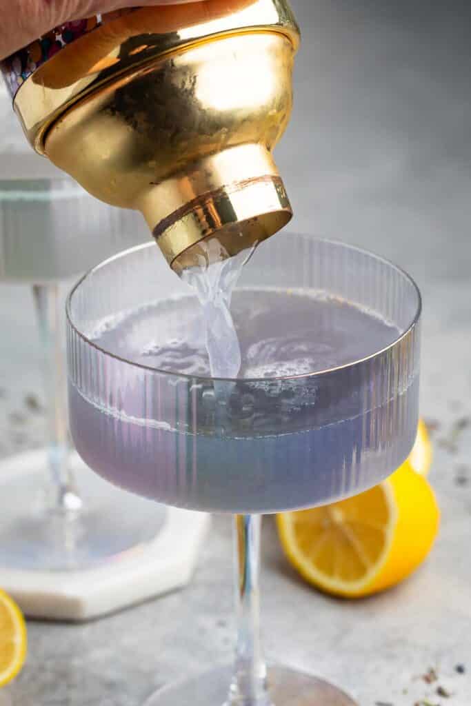 A gold cocktail shaker pouring a clear liquid into a ribbed glass cup. Two lemon halves and another similar glass are visible in the background.