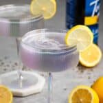 Two iridescent cocktail glasses filled with a violet-hued drink, each garnished with a lemon slice. Whole and halved lemons are scattered on the gray surface. A bottle is blurred in the background.