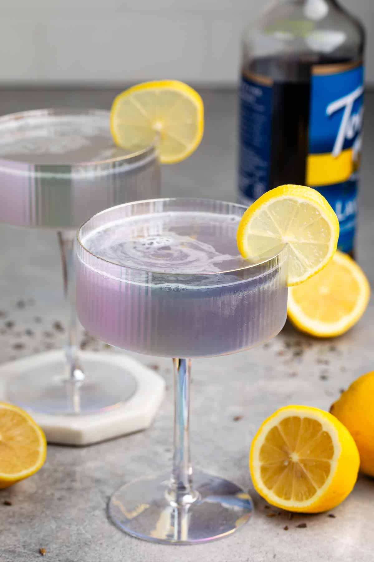 Two iridescent cocktail glasses filled with a violet-hued drink, each garnished with a lemon slice. Whole and halved lemons are scattered on the gray surface. A bottle is blurred in the background.