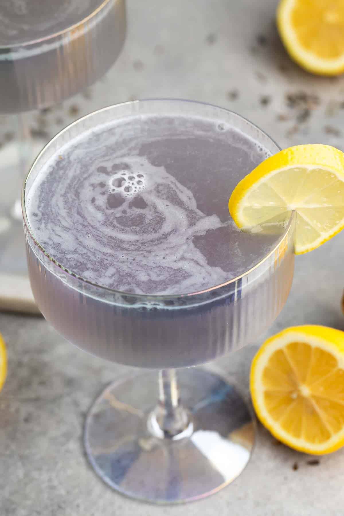 A cocktail glass filled with a lavender-colored drink, garnished with a lemon slice on the rim. Lemon halves are scattered on the gray surface around the glass.