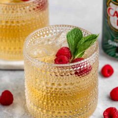 Two textured glasses filled with a light brown beverage, garnished with raspberries and mint leaves. Fresh raspberries and sprigs of mint are scattered on the surface. A green can is partially visible in the background.