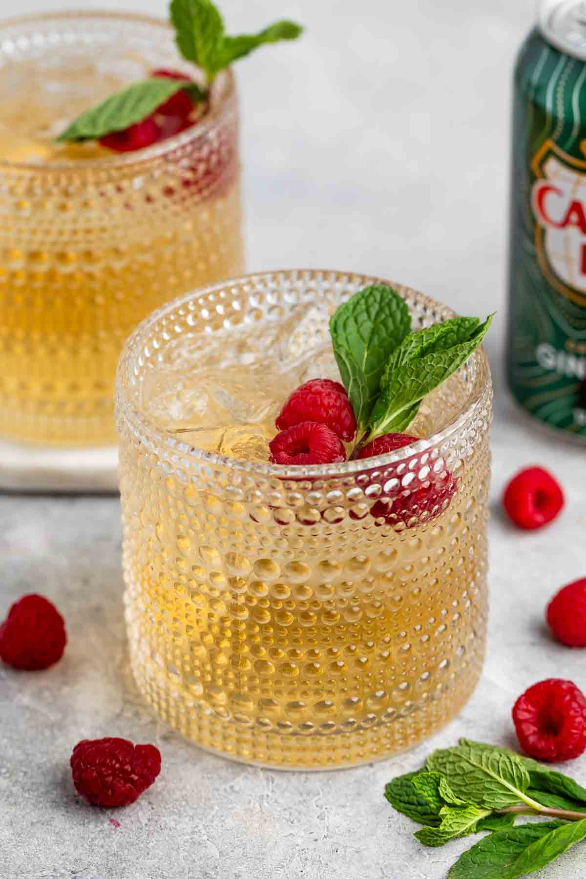 Two textured glasses filled with a light brown beverage, garnished with raspberries and mint leaves. Fresh raspberries and sprigs of mint are scattered on the surface. A green can is partially visible in the background.