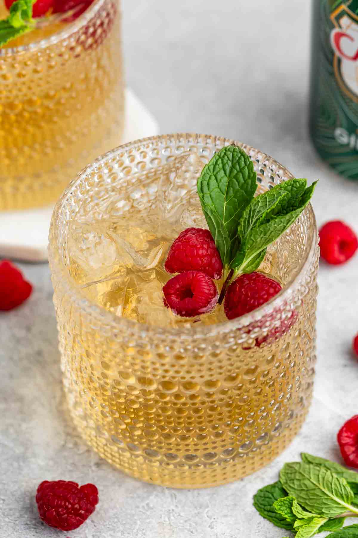 A textured glass filled with a light golden drink topped with ice, fresh raspberries, and mint leaves. More raspberries and mint leaves are scattered around the glass on a light gray surface. Another similar drink is partially visible in the background.