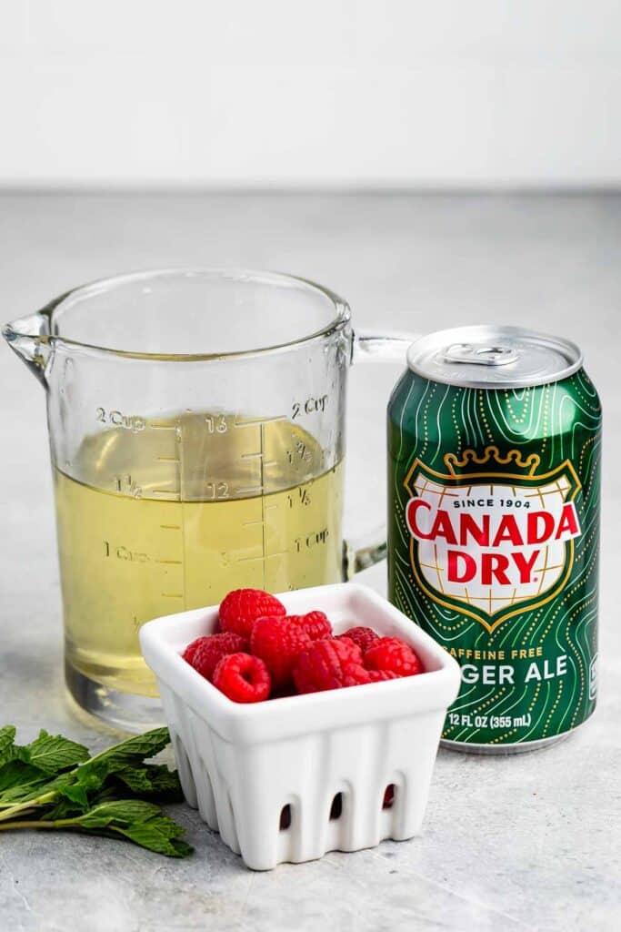 A can of Canada Dry ginger ale is next to a measuring cup filled with liquid, a white container of raspberries, and a few sprigs of mint on a light gray surface.