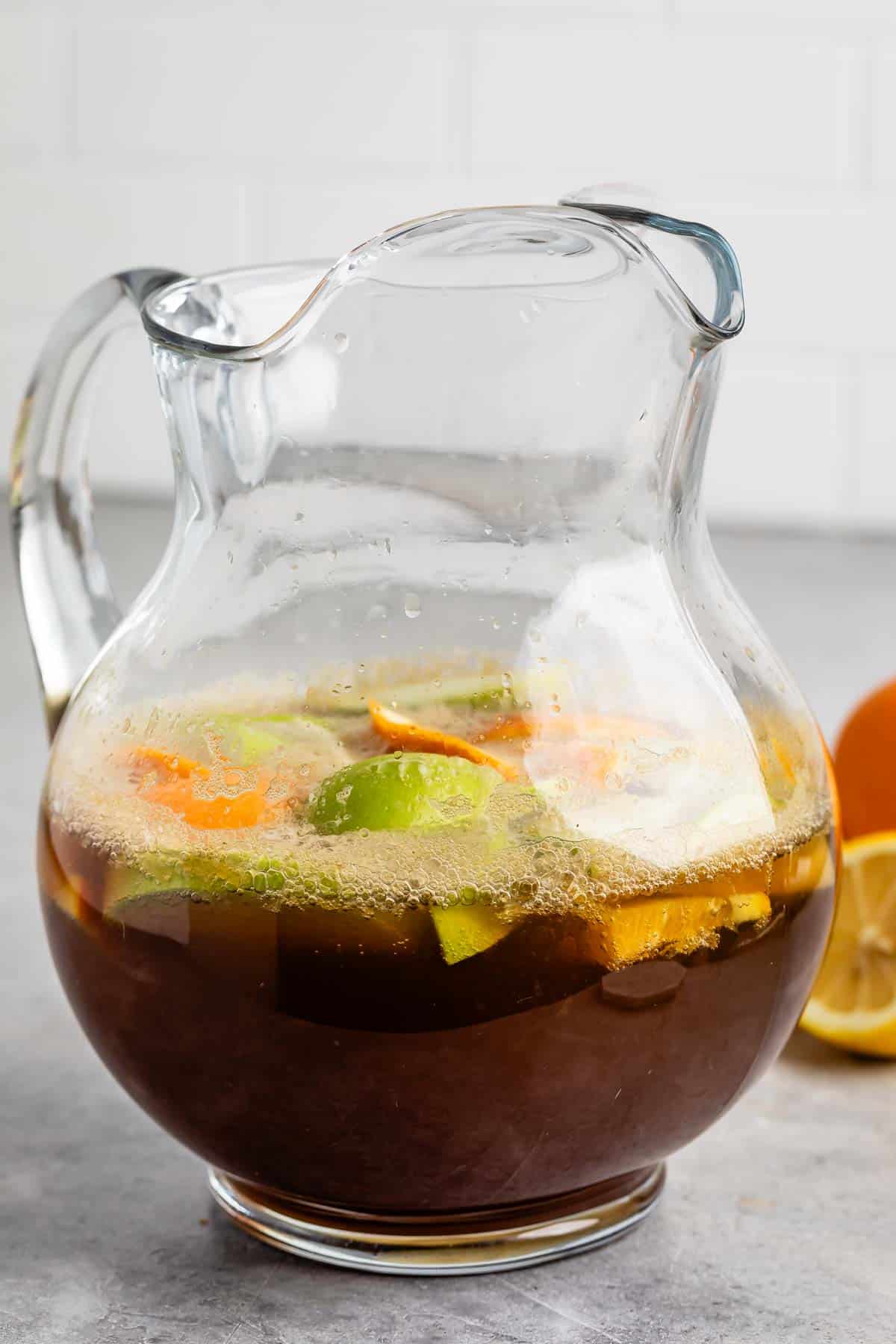 A glass pitcher filled with a dark beverage, containing visible slices of green lime, orange, and lemon. The pitcher is placed on a gray countertop with a blurred background.