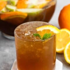 A chilled, brown drink in a glass with ice, garnished with an orange slice and mint. Behind it, a pitcher filled with mixed citrus fruits and liquid. Orange and lemon slices are placed nearby on a gray surface.