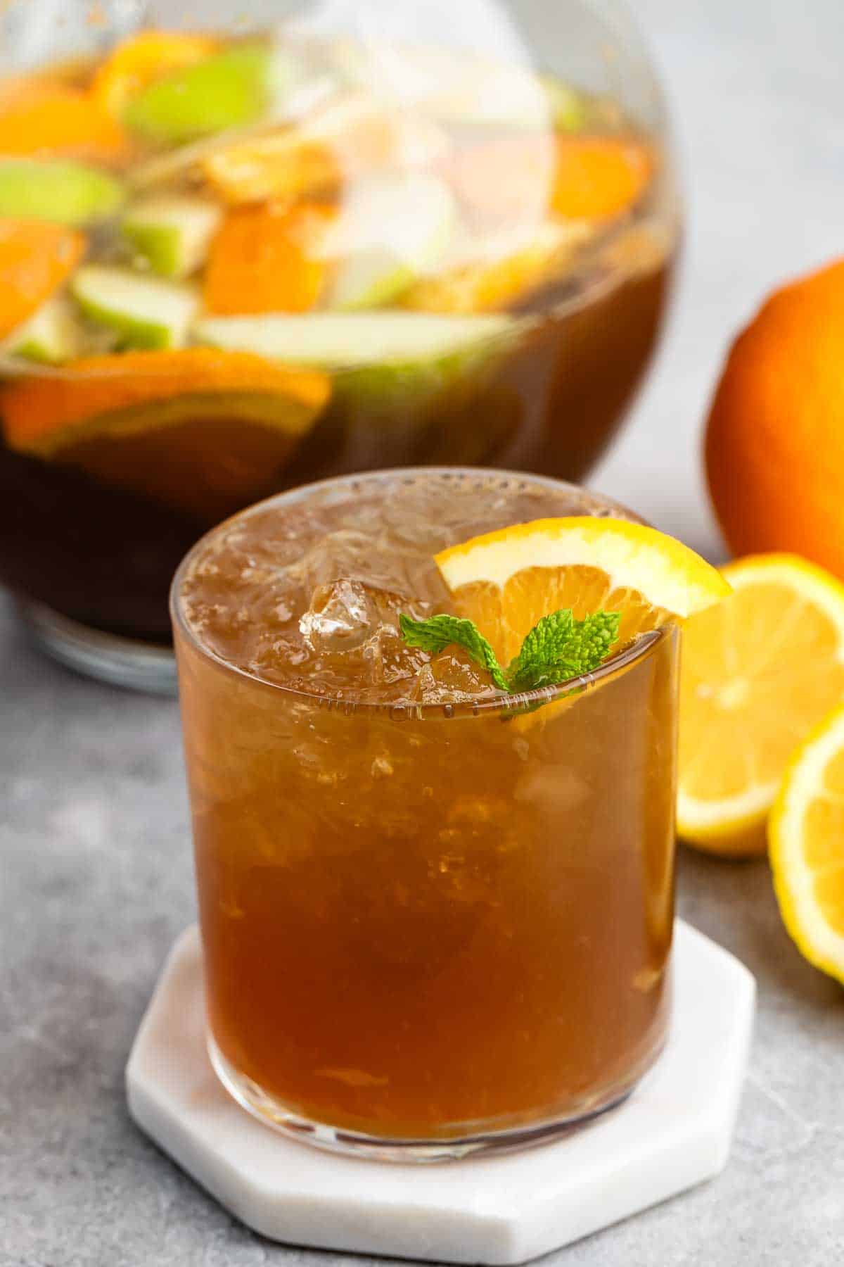 A chilled, brown drink in a glass with ice, garnished with an orange slice and mint. Behind it, a pitcher filled with mixed citrus fruits and liquid. Orange and lemon slices are placed nearby on a gray surface.