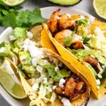 Two grilled shrimp tacos on a plate with corn tortillas, topped with avocado slices, lettuce, cilantro, and cream. Lime wedges and an avocado half are beside the plate, adding freshness to the presentation.