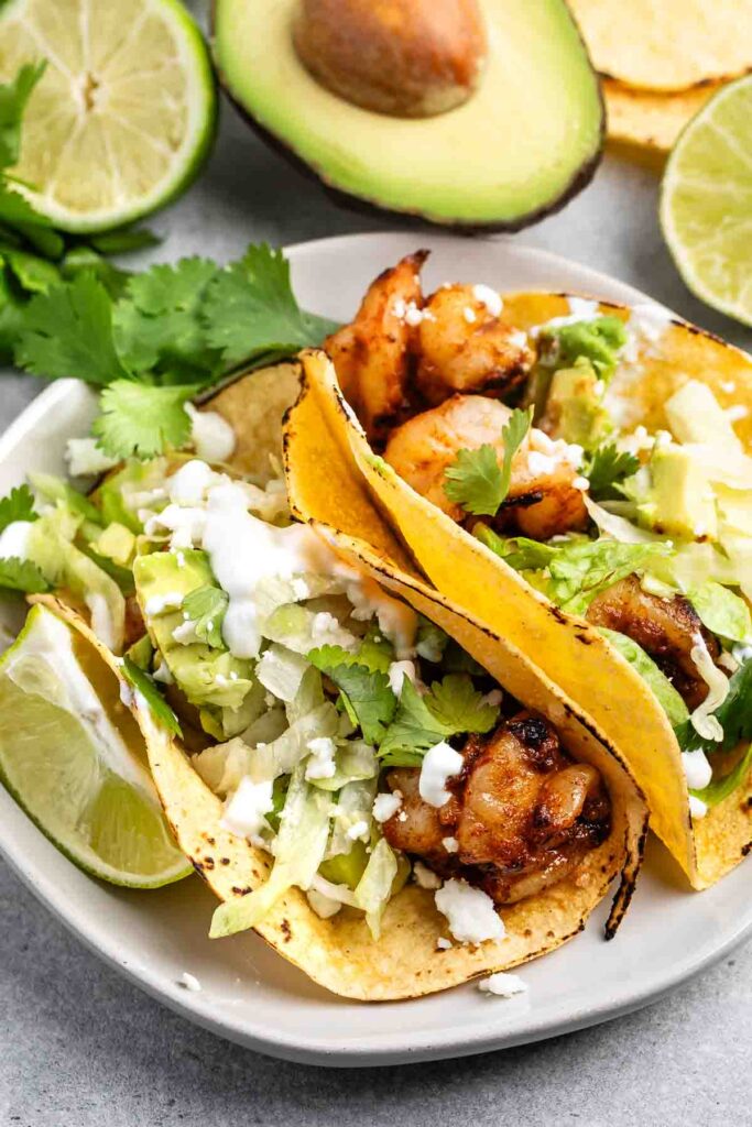 Two grilled shrimp tacos on a plate with corn tortillas, topped with avocado slices, lettuce, cilantro, and cream. Lime wedges and an avocado half are beside the plate, adding freshness to the presentation.