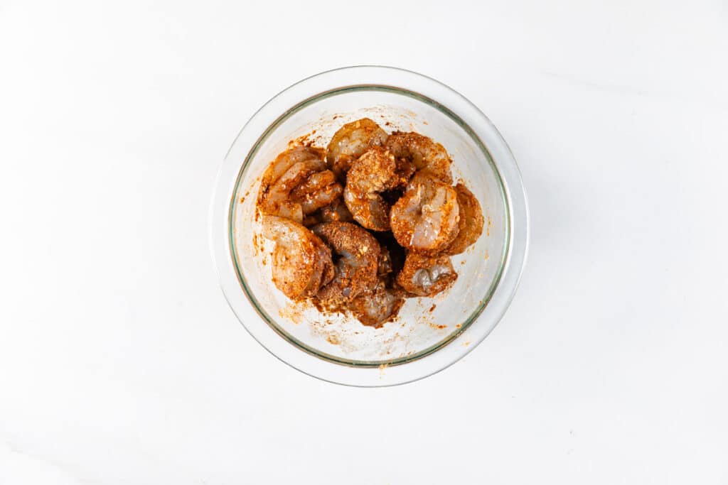 Glass bowl containing raw shrimp coated in a seasoned marinade, placed on a white surface.