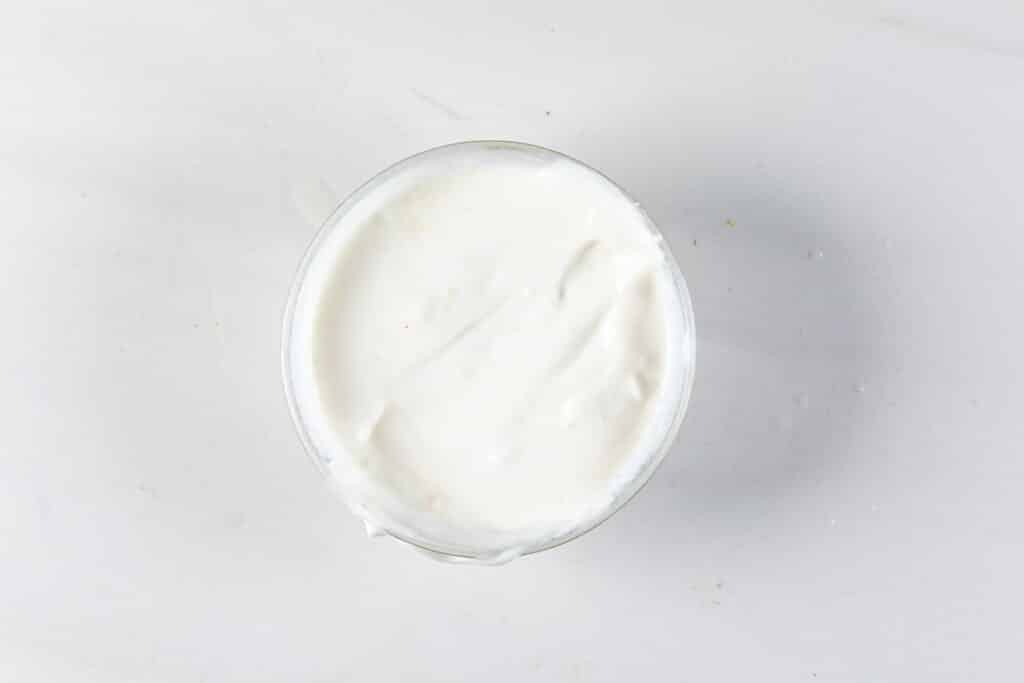 A glass bowl filled with smooth, white yogurt sits on a light gray surface. The texture is creamy and consistent, with soft shadows surrounding the bowl.