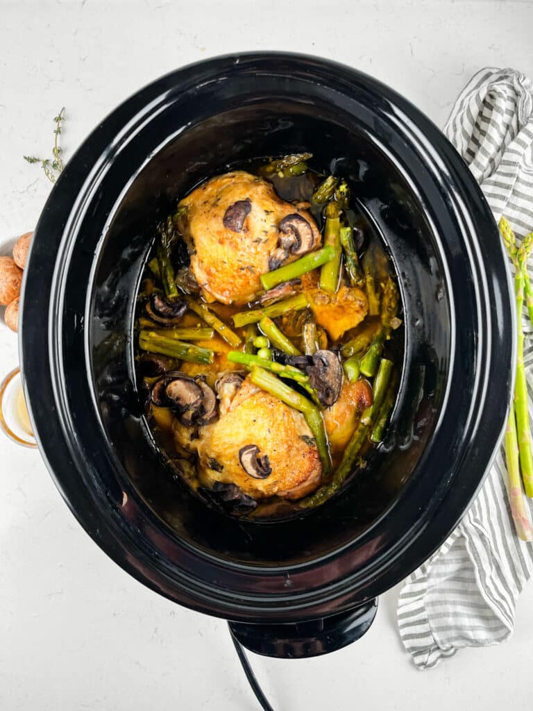 A slow cooker filled with cooked chicken thighs, mushrooms, asparagus, and sliced vegetables. The dish is surrounded by a gray-striped cloth and fresh asparagus on a white countertop.