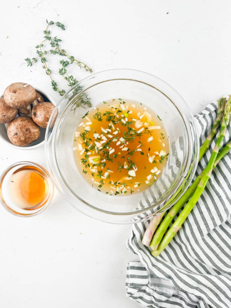 A glass bowl filled with a brown marinade containing chopped onions and herbs. Next to it are fresh thyme, mushrooms on a small dish, a bunch of asparagus, a striped cloth, and a small glass bowl of brown liquid on a white surface.