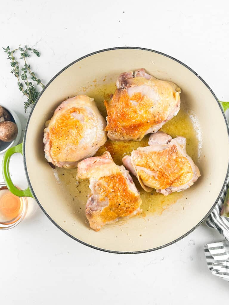 Four golden-brown chicken pieces are cooking in a round, green-edged pot. Fresh herbs are nearby, along with a small container of spices, on a white surface. A striped cloth is partially visible on the right.