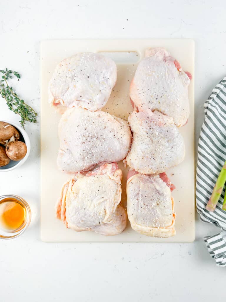 Raw seasoned chicken thighs on a cutting board, sprinkled with salt and pepper. A bowl of mushrooms, a sprig of thyme, a glass of orange liquid, and a striped cloth are nearby on a light countertop.