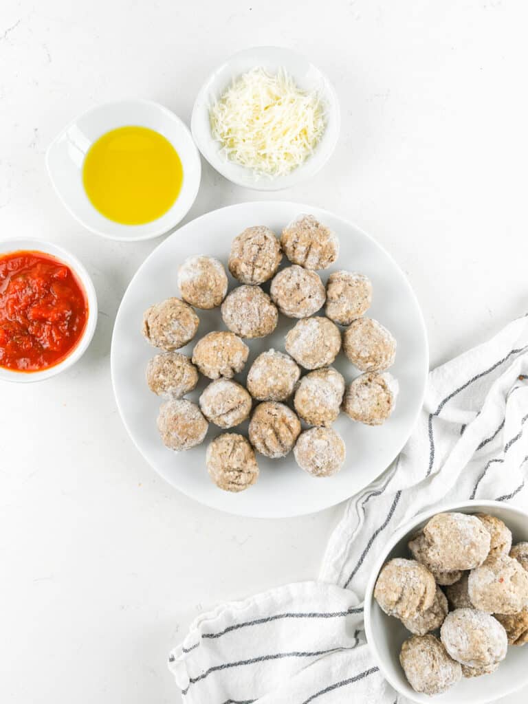 A plate of uncooked meatballs is surrounded by bowls containing shredded cheese, tomato sauce, and olive oil on a white surface. Another bowl of meatballs and a striped cloth napkin are nearby.