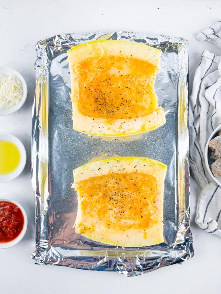 Two halves of a spaghetti squash, seasoned with salt and pepper, are placed cut side up on a foil-lined baking sheet. Surrounding them are small bowls of olive oil, grated cheese, marinara sauce, and a striped towel.