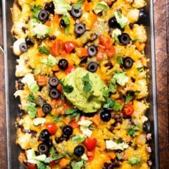 A tray of loaded tater tot nachos topped with melted cheese, black olives, cherry tomatoes, avocado, cilantro, and a large dollop of guacamole on a wooden table.