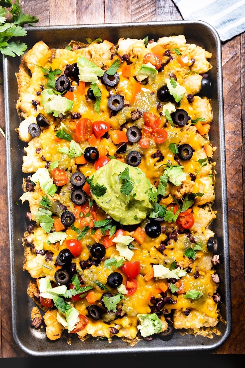 A tray of loaded tater tot nachos topped with melted cheese, black olives, cherry tomatoes, avocado, cilantro, and a large dollop of guacamole on a wooden table.