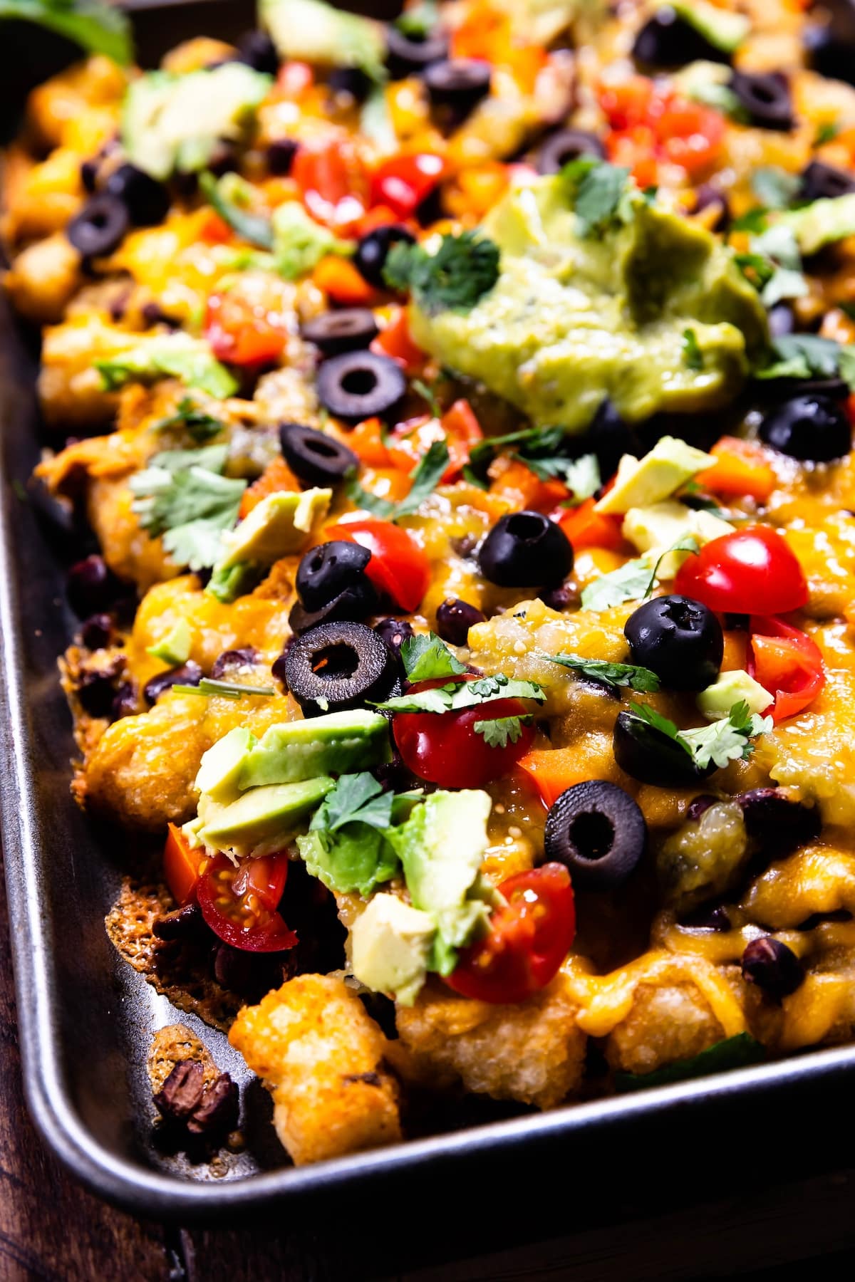 A close-up of a tray filled with loaded tater tots topped with melted cheese, sliced black olives, cherry tomatoes, diced avocado, cilantro, and a dollop of guacamole.