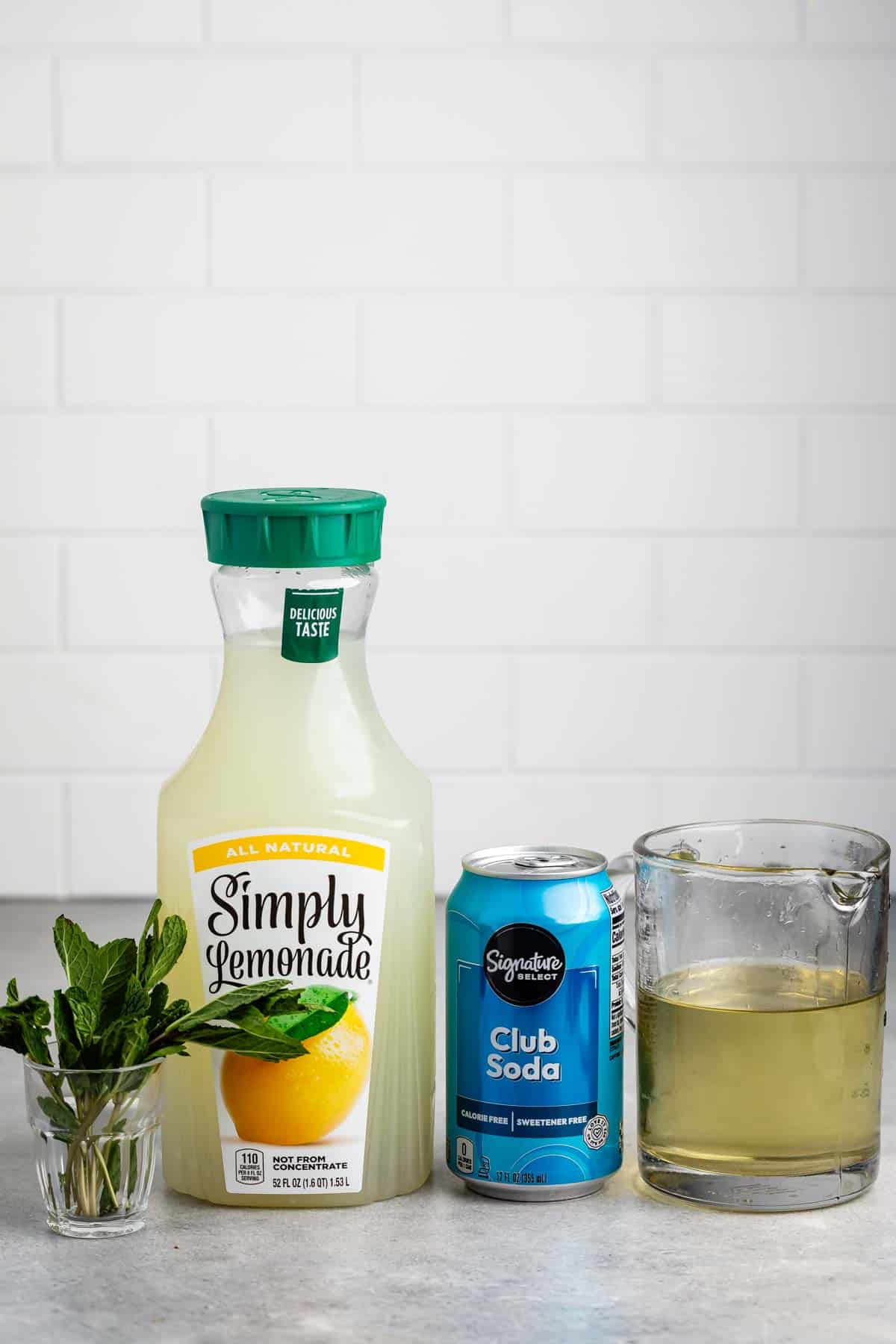 A bottle of Simply Lemonade, a can of club soda, a measuring cup with liquid, and a small glass with fresh mint leaves are arranged on a light surface. The background features a white tiled wall.