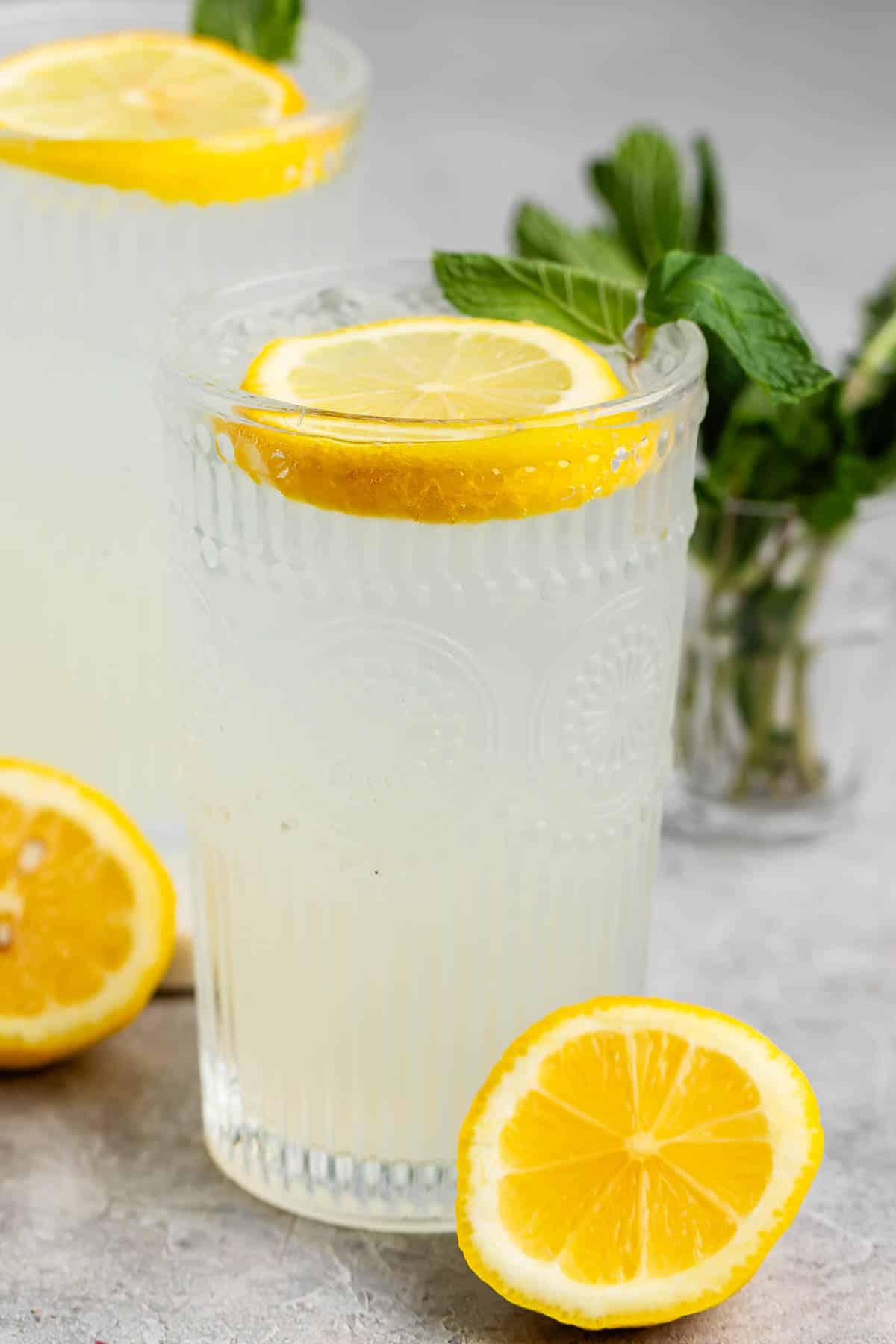 A glass of lemonade with a lemon slice and mint sprig on top sits on a gray surface. Another glass and a bunch of mint leaves are in the background. Sliced lemons are placed next to the glass.