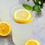 A refreshing glass of lemonade with a slice of lemon floating on top. A sprig of mint is placed at the rim. A whole lemon and another glass of lemonade are in the background on a light, textured surface.