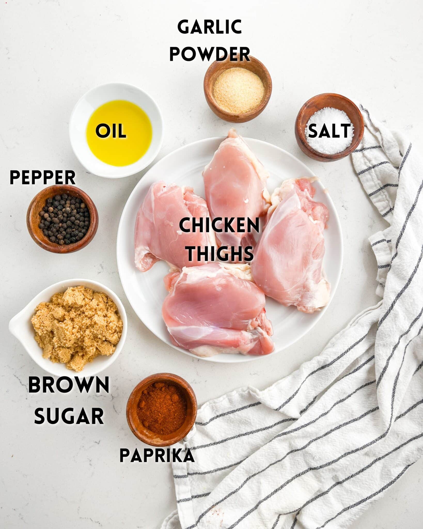 A flat lay of ingredients for cooking includes raw chicken thighs on a plate, surrounded by small bowls labeled with salt, pepper, brown sugar, paprika, garlic powder, and oil. A striped cloth lies beside the arrangement.