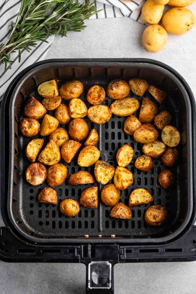 Golden roasted potatoes in an air fryer basket, garnished with herbs. Fresh rosemary and a bunch of small raw potatoes are nearby on a gray countertop.