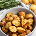 A bowl of seasoned roasted potatoes sits on a countertop. Fresh rosemary is visible in the background alongside whole potatoes. The potatoes are golden brown and appear crispy, suggesting a savory and flavorful dish.