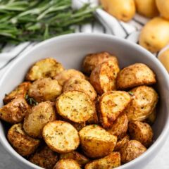 A bowl of seasoned roasted potatoes sits on a countertop. Fresh rosemary is visible in the background alongside whole potatoes. The potatoes are golden brown and appear crispy, suggesting a savory and flavorful dish.