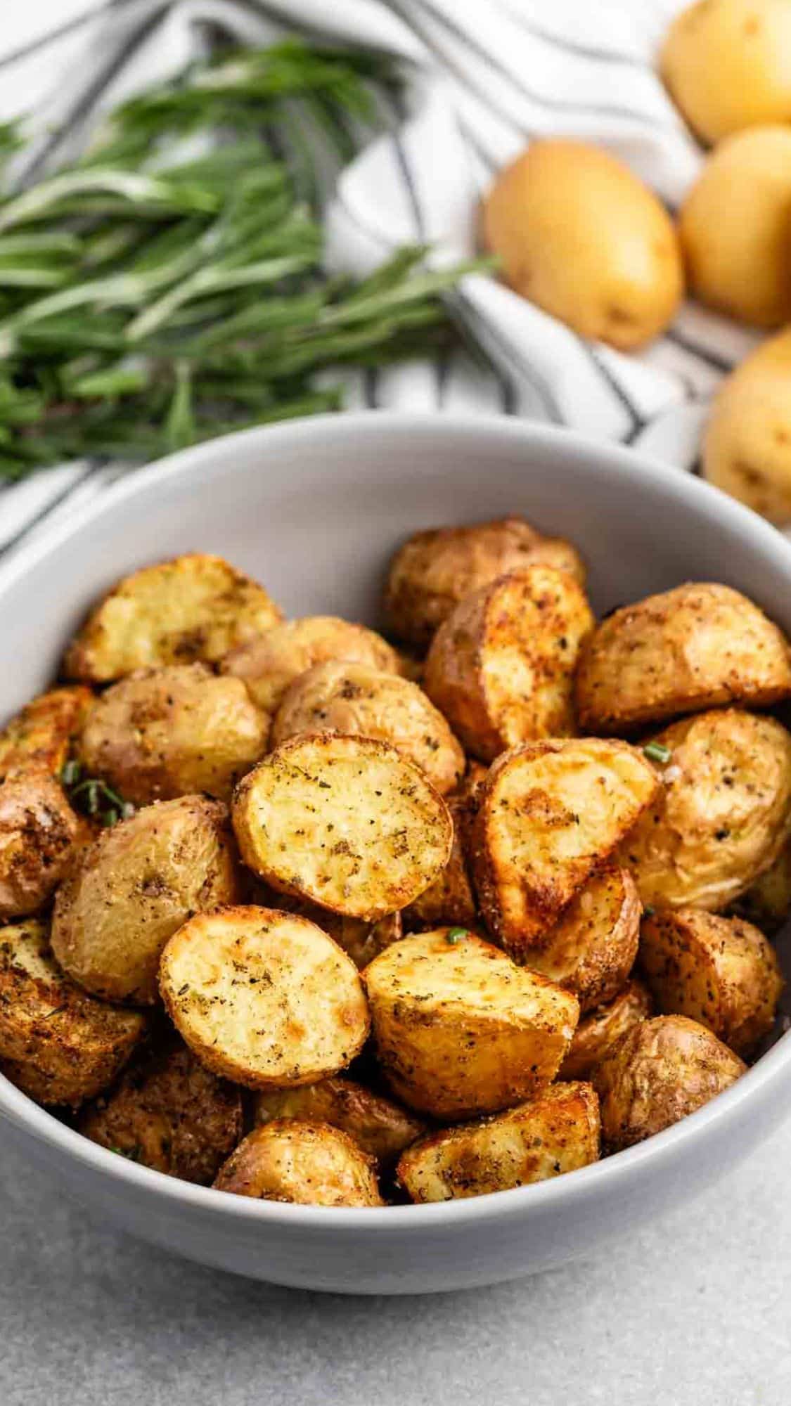 A white bowl filled with seasoned roasted potato halves, positioned on a light-colored surface. Fresh rosemary and whole potatoes are in the background, along with a striped cloth.